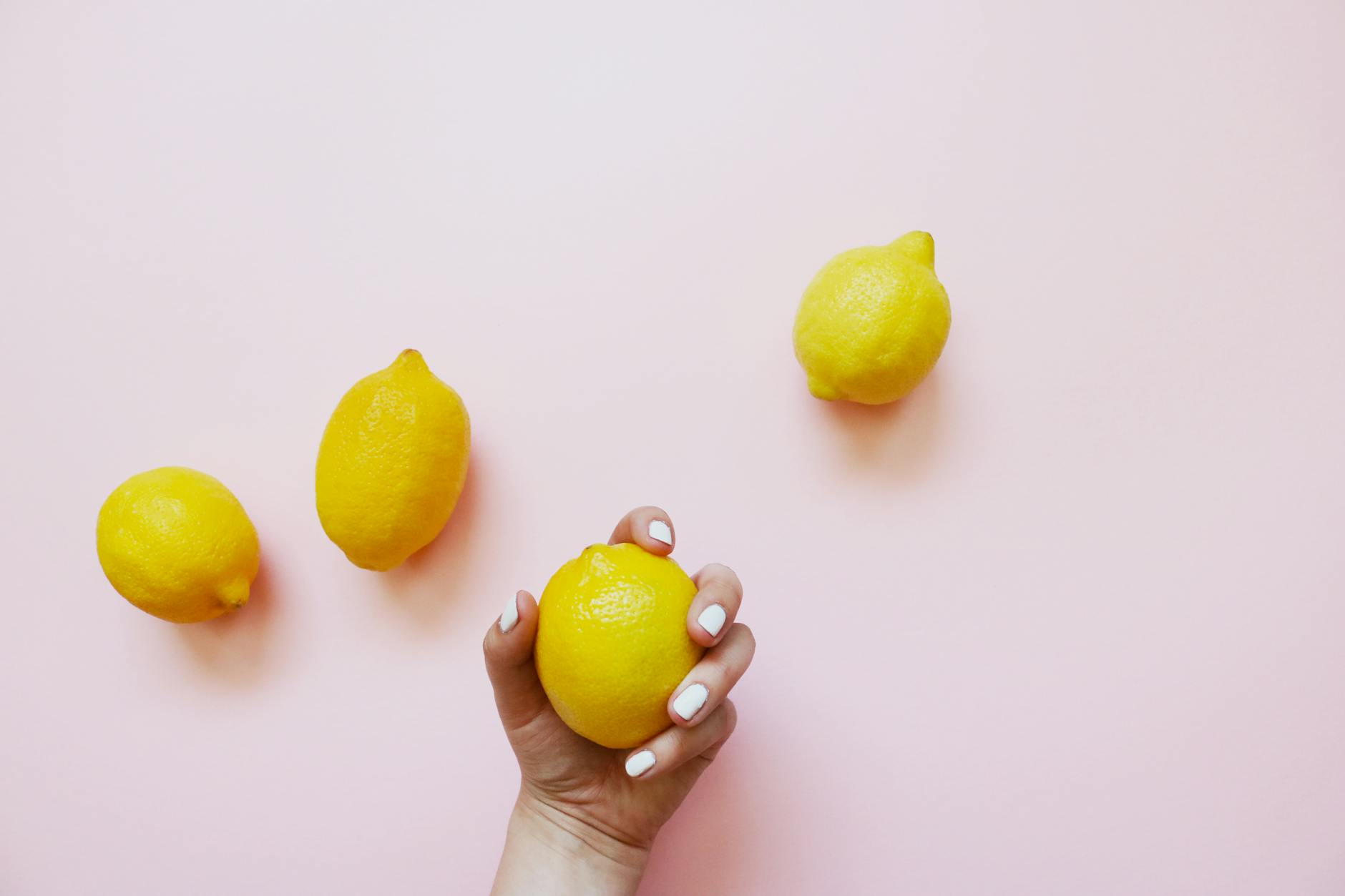 Mano con uñas blancas sosteniendo un limón sobre fondo rosa suave, rodeado de tres limones adicionales