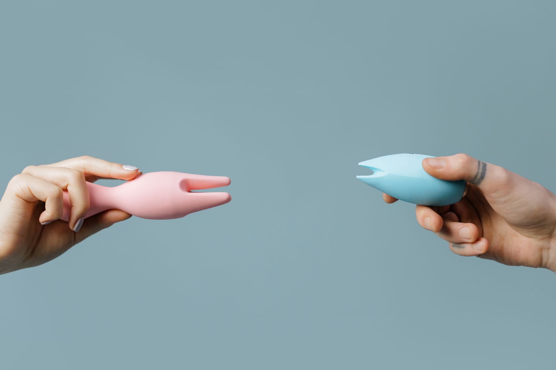Two hands holding pink and blue silicone vibrators together against a pastel background